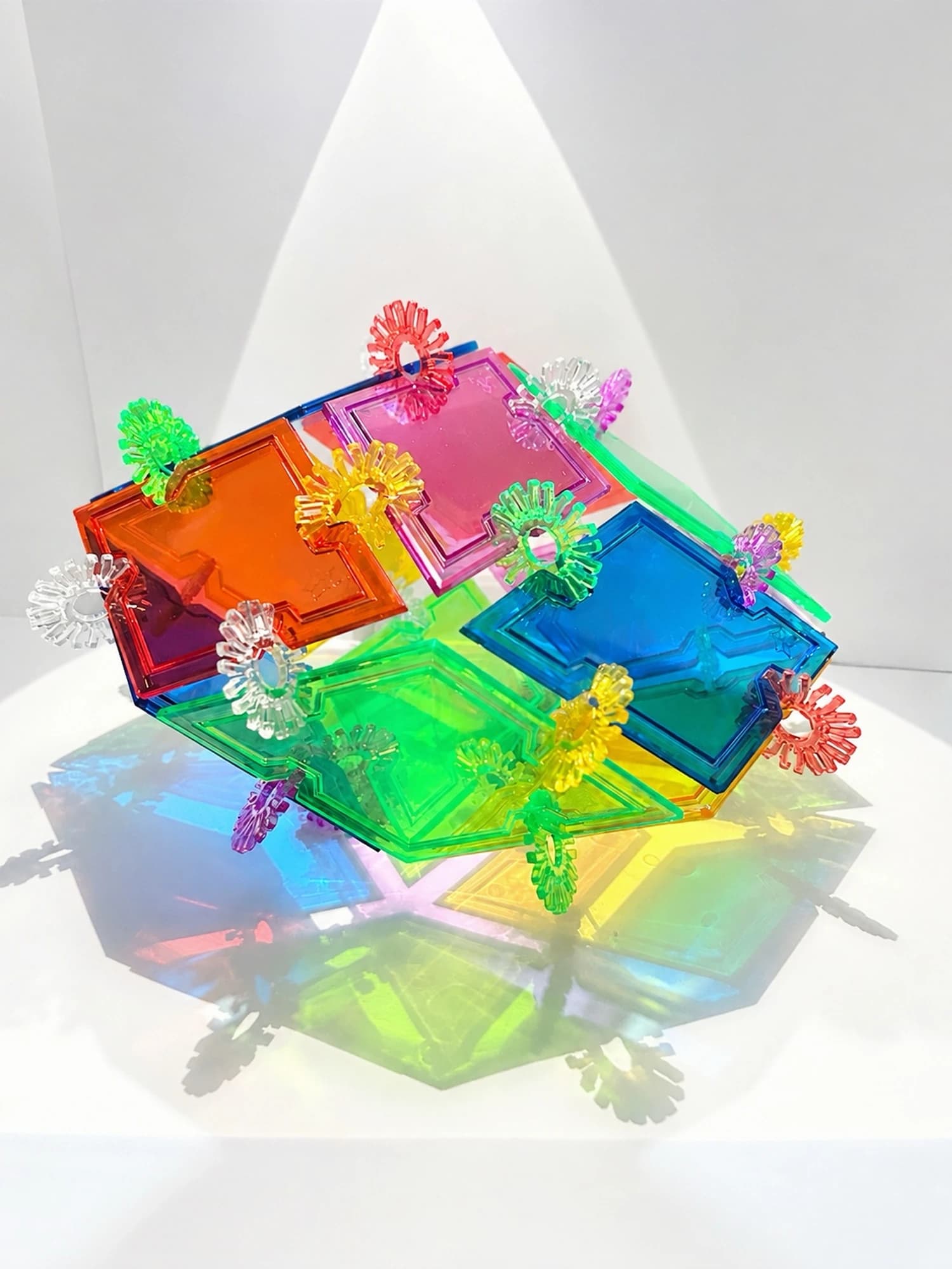 A low polyhedron with orange, pink, blue and green rhombus faces casting colourful shadows.